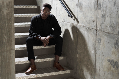 Menswear shot of a man in all black seated on concrete steps. By Nick van der Touw.