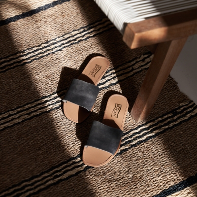 Product shot of black suede slide sandals on a woven rug. By Nick van der Touw.