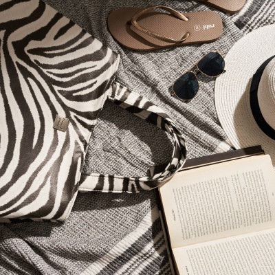 Beach lifestyle flat lay with a straw hat, zebra-print bag and book. By Nick van der Touw.