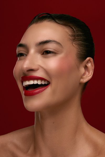 Beauty profile portrait of Albina with a red lip mark on the cheek, red backdrop. Studio editorial by Nick van der Touw.